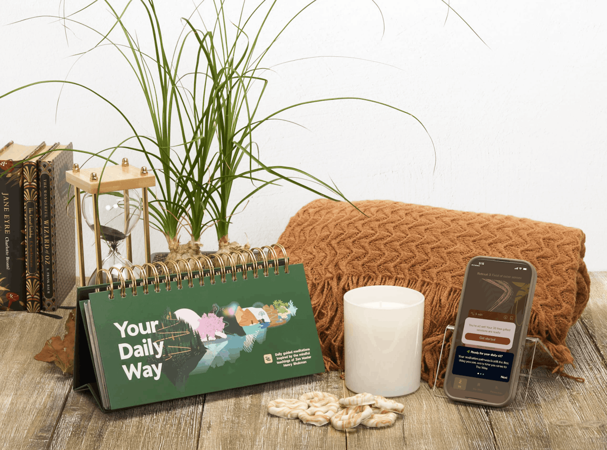 "Your Daily Way" table top calendar with candle, brown folded blanket, hourglass, plant, and books
