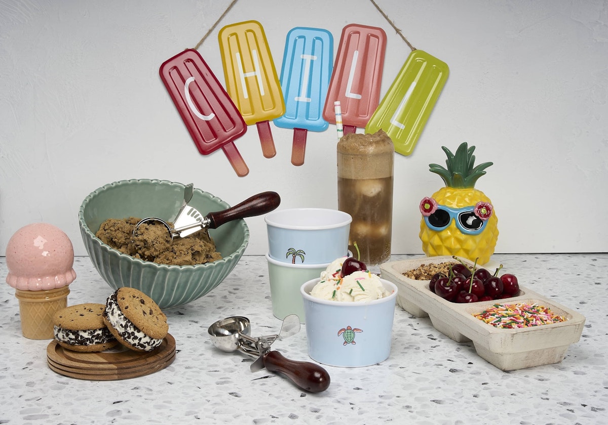 Sign made of popsicles that spell the word 'CHILL', ice cream cookie sandwiches, green bowl with scoop scooping out cookie dough, ice cream cups and setup station with toppings including sprinkles, cherries, and nuts, root beer float in clear glass, and ceramic pineapple with sunglasses.