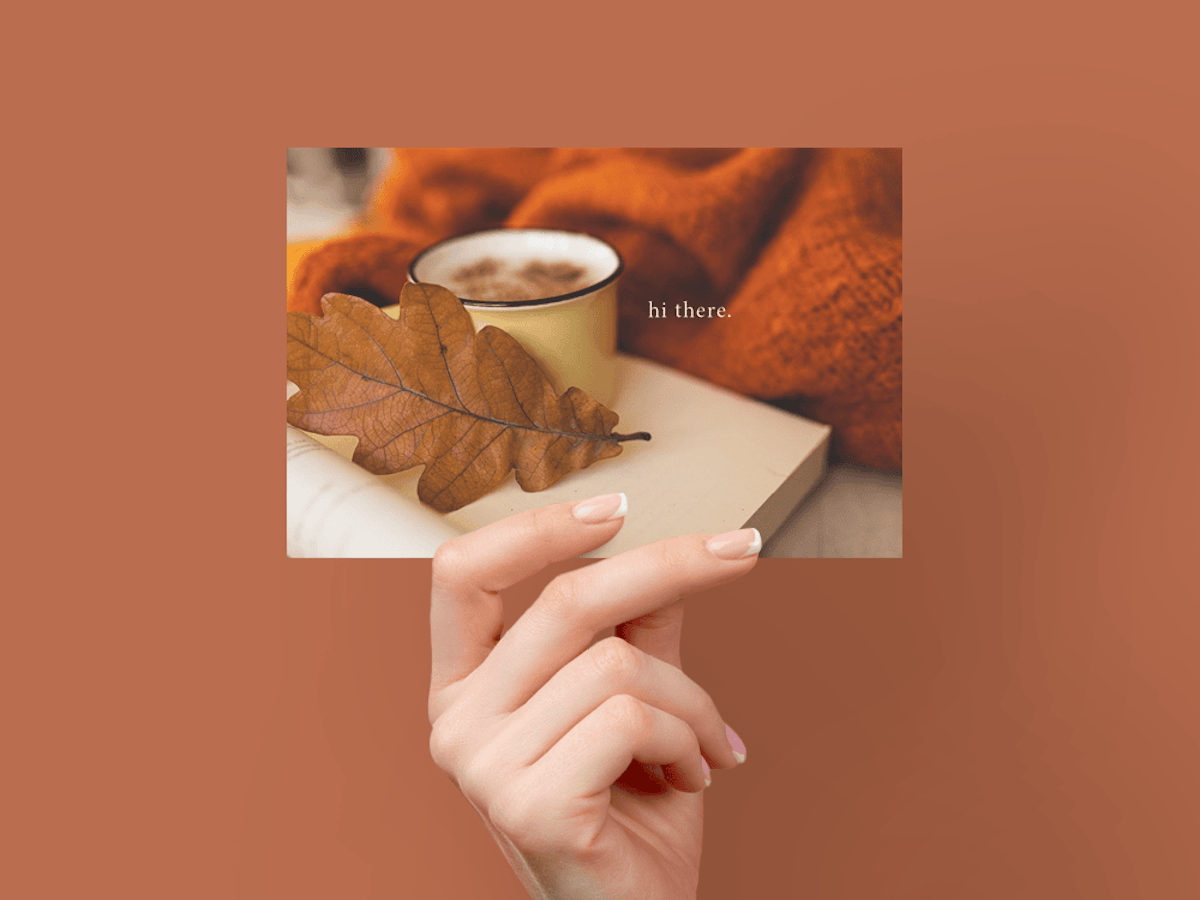 Hand holding card with fall leaf, cup with hot beverage, orange blanket, and words 'hi there.'